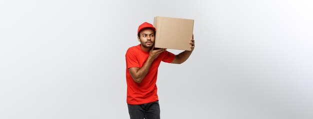 Concept de livraison Portrait d'un livreur afro-américain curieux écouter à l'intérieur d'un paquet de boîte isolé sur fond de studio gris espace de copie