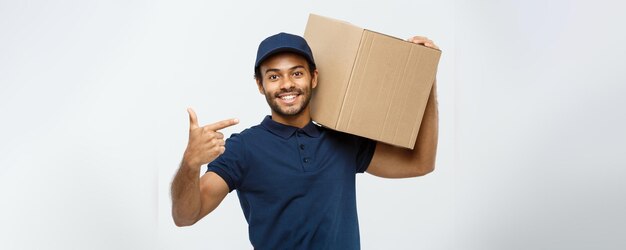 Concept de livraison Portrait d'un livreur afro-américain heureux pointant la main pour présenter un colis isolé sur fond gris studio Espace de copie