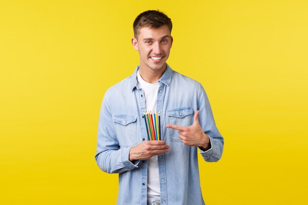 Concept de loisirs, de loisirs et de mode de vie des personnes. Beau jeune homme souriant, artiste pointant du doigt des crayons de couleur de bonne qualité, fond jaune debout recommandant un produit de dessin