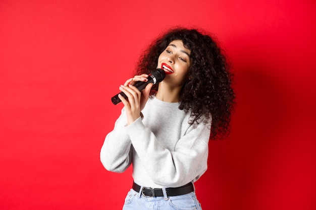 Concept de loisirs et de passe-temps. Jeune femme élégante chantant un karaoké, regardant de côté et tenant un microphone, interprétant une chanson, debout sur fond rouge