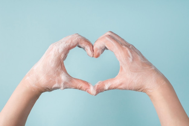 Concept de lutte contre le coronavirus. photo recadrée en gros plan de mains féminines montrant la forme de coeur avec deux mains dans de la mousse de gel de savon sur fond de couleur bleu pastel