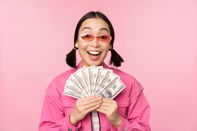 Concept de microcrédit et de prêts rapides Fille coréenne élégante et excitée montrant de l'argent en dollars et ayant l'air heureux debout dans des lunettes de soleil sur fond rose