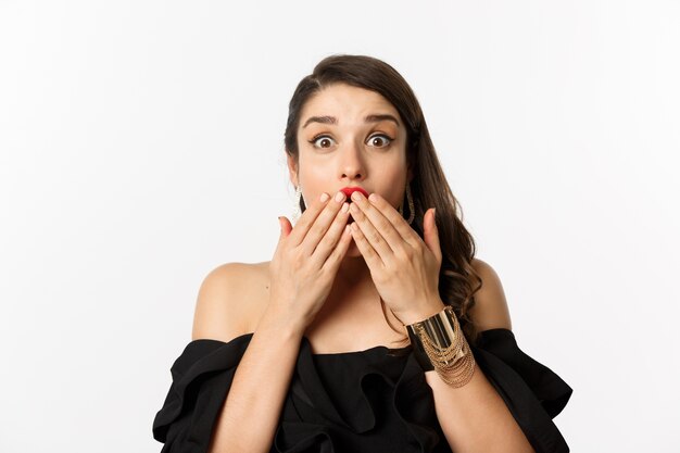 Concept de mode et de beauté. Gros plan d'une jeune femme excitée à la surprise, couvrir la bouche avec les mains et regarder la caméra avec joie, fond blanc.