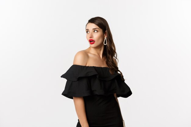 Concept de mode et de beauté. Image de la jeune femme en robe noire tourner derrière et en regardant l'espace de copie, debout sur fond blanc.