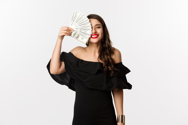 Concept de mode et de shopping. Heureuse jeune femme en robe noire, avec des lèvres rouges, tenant de l'argent et souriant satisfait, debout sur fond blanc.