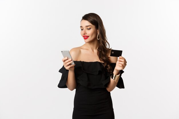 Concept de mode et de shopping. Jeune femme séduisante faisant des achats en ligne, achetant sur internet avec carte de crédit et smartphone, fond blanc.