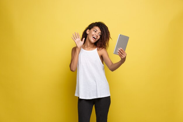 Concept de mode de vie - Portrait de la belle femme afro-américaine joyeuse jouant quelque chose sur la tablette électronique. Contexte en pastel pastel jaune. Espace de copie.