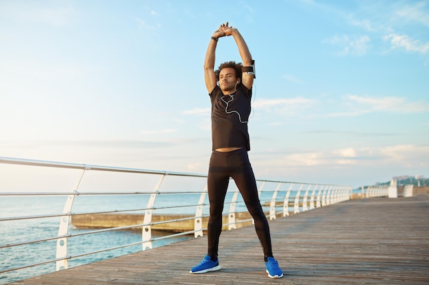 Concept de mode de vie sain Garçon sportif à la peau foncée en vêtements de sport noirs et écouteurs debout sur une plate-forme en bois