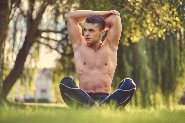 Concept de mode de vie sain. Jeune homme torse nu qui s'étire les mains tout en étant assis en posture de lotus sur l'herbe verte.