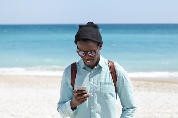 Concept de mode de vie, de technologie et de communication moderne. Séduisante jeune hipster européenne noire en chemise, chapeau et lunettes de soleil bénéficiant d'une connexion Internet haute vitesse 3g ou 4g tout en passant un week-end au bord de la mer