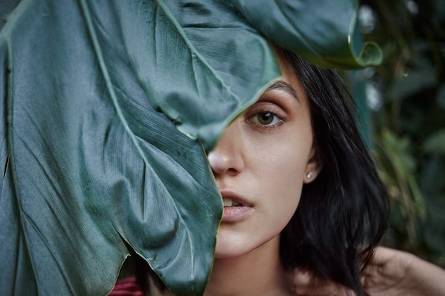 Photo gratuite concept de nature, beauté, soins de la peau et cosmétiques bio. la moitié du visage bouchent le portrait de la magnifique jeune femme aux cheveux bob avec une peau propre posant à l'extérieur, se cachant derrière une grande feuille verte fraîche