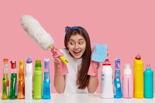 Concept de nettoyage de printemps. Satisfait brunette jeune femme de race blanche tient un pinceau à poussière et une éponge bleue, sourit positivement