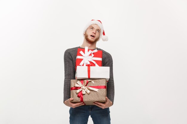 Concept de Noël jeune bel homme avec barbe tenant des cadeaux lourds avec une expression faciale épuisée sur fond blanc