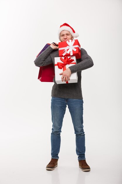 Concept de Noël jeune bel homme avec barbe tenant des cadeaux lourds et des sacs à provisions avec une expression faciale épuisée sur fond blanc