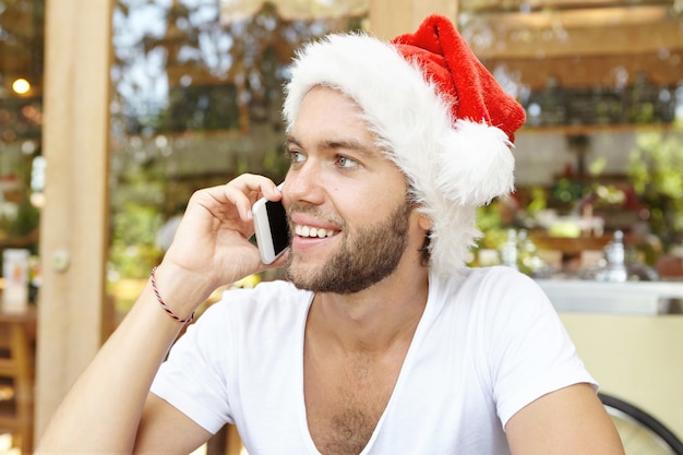 Concept de Noël et de joyeuses fêtes. Séduisante jeune homme avec une barbe élégante portant un t-shirt blanc et un chapeau de père Noël parlant au téléphone mobile