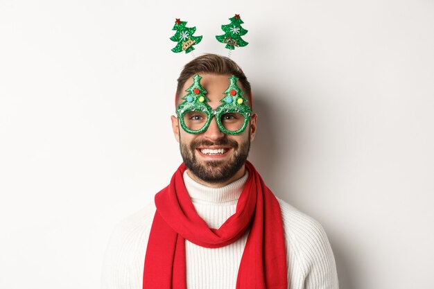 Concept de Noël, nouvel an et célébration. Gros plan d'un bel homme barbu dans des lunettes de fête amusantes à l'air heureux, debout sur fond blanc