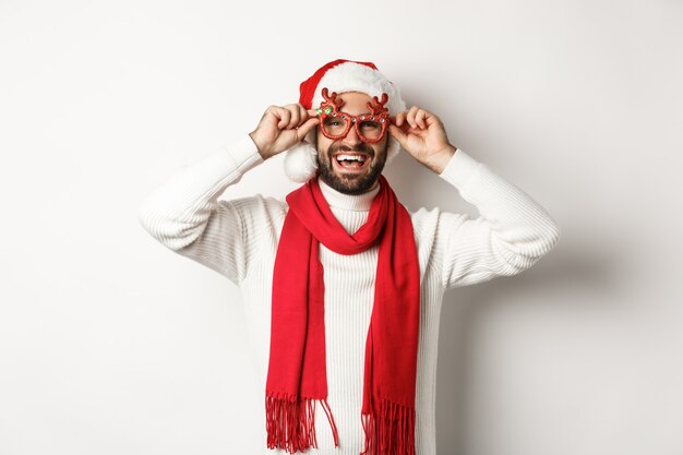 Concept de Noël, nouvel an et célébration. Heureux homme riant, portant un bonnet de Noel et des lunettes de fête, debout sur fond blanc