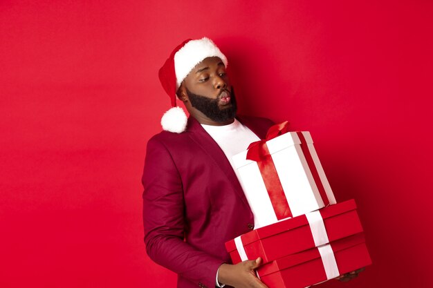 Concept de Noël, nouvel an et shopping. Funny african american man in santa hat porter de lourds cadeaux de Noël, tenant des cadeaux, debout sur fond rouge