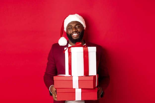Concept de noël, nouvel an et shopping. heureux homme noir en bonnet de noel et blazer tenant des cadeaux de noël, apporte des cadeaux et souriant, debout sur fond rouge.