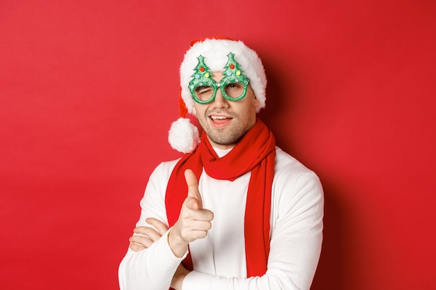 Concept de noël, vacances d'hiver et célébration. Gros plan d'un jeune homme effronté en bonnet de noel et lunettes de fête, souriant et pointant le pistolet à la caméra, debout sur fond rouge.