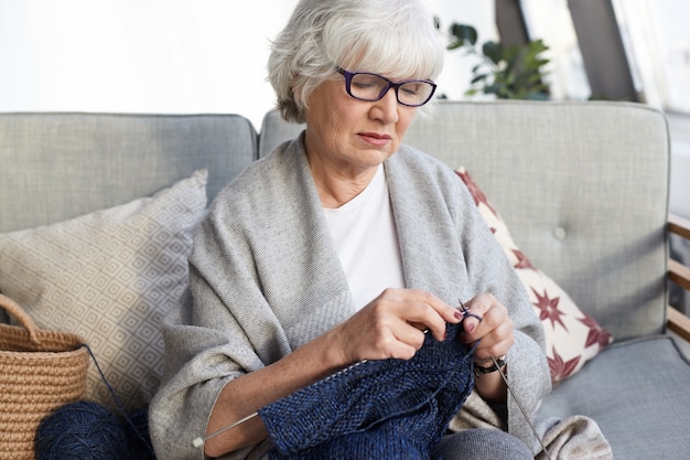 Concept de passe-temps, de loisirs et de retraite. Beau grand-mère élégante portant des lunettes assis sur un canapé gris avec des aiguilles, pull à tricoter pour son petit-fils, ayant un regard sérieux et concentré