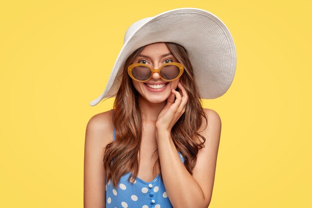 Concept de personnes, de beauté et de style. Contenu joyeuse femme européenne porte un chapeau blanc, une robe à pois, des lunettes de soleil, sourit largement, a de la joie comme recrée dans un lieu de villégiature, isolé sur un mur jaune.