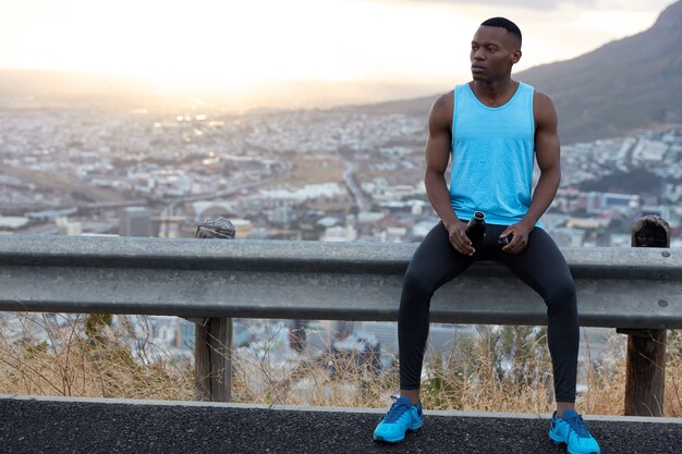 Concept de personnes, de bien-être et d'ethnicité. Photo d'un homme à la peau foncée en tenue de sport, porte une bouteille avec de l'eau, s'assoit au panneau de signalisation, des modèles sur la nature avec un espace libre pour votre annonce