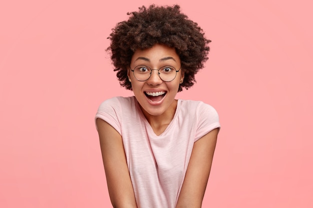 Photo gratuite concept de personnes, d'émotions et de sentiments agréables. belle jeune femme afro-américaine avec une expression joyeuse, sourit joyeusement en regardant le programme drôle pendant le temps libre, porte un t-shirt décontracté