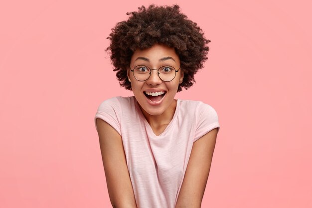 Concept de personnes, d'émotions et de sentiments agréables. Belle jeune femme afro-américaine avec une expression joyeuse, sourit joyeusement en regardant le programme drôle pendant le temps libre, porte un t-shirt décontracté