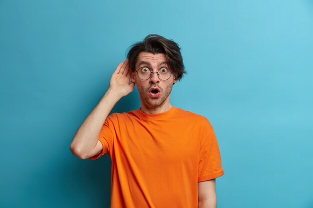 Concept de personnes, d'émotions et de style de vie. Surpris, un homme étonné perd la parole de l'émerveillement, le regarde avec les yeux écarquillés et la bouche ouverte, entend de terribles nouvelles, porte un t-shirt orange, des lunettes transparentes