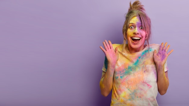 Concept de personnes, d'émotions et de vacances. Une femme européenne heureuse et émotive soulève des paumes de bonheur, ne peut pas arrêter les sentiments positifs, étant impressionnée par la célébration du Festival des couleurs de Holi à l'étranger