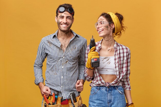 Photo gratuite concept de personnes, emploi, profession, profession, travail d'équipe et succès. équipe de personnel de maintenance bénéficiant des résultats de leur travail commun: belle femme avec une perceuse regardant l'homme et souriant joyeusement