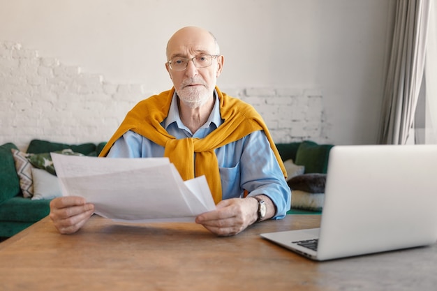 Concept de personnes, finances, technologie et emploi. Sérieux homme d'affaires à la retraite à la mode faisant des finances au bureau moderne, tenant des papiers dans ses mains, ordinateur portable ouvert sur une table en bois à partir de lui