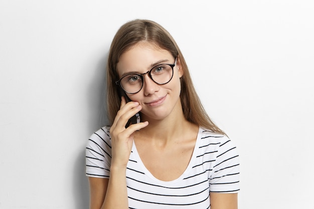 Concept de personnes et de gadgets électroniques modernes. Adolescente européenne mignonne en t-shirt rayé et lunettes profitant d'une conversation téléphonique avec son meilleur ami, discutant des garçons, des potins et des devoirs