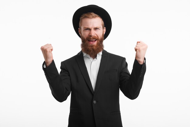 Concept de personnes, de joie, de bonheur et de succès. Heureux jeune homme d'affaires aux cheveux roux barbu confiant portant rond élégant avait et costume s'exclamant de manière gagnante et avec enthousiasme, levant les poings fermés