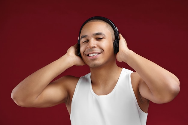 Concept de personnes, de joie, de détente, de divertissement et de technologie moderne. Image studio isolé de joyeux gars africain positif portant un débardeur blanc, bénéficiant d'une bonne musique dans de nouveaux écouteurs sans fil