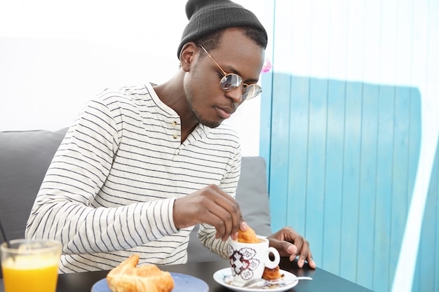 Concept De Personnes, De Nourriture, De Loisirs Et De Style De Vie. Beau Mec Afro-américain à La Mode Dans Des Lunettes à La Mode Et Des Couvre-chefs Trempant Un Croissant Dans Une Tasse De Café Tout En Dégustant Un Délicieux Petit Déjeuner Au Café