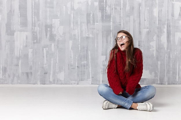 Concept de personnes, de repos et de relaxation. Belle jeune femme caucasienne joyeuse en tenue élégante assise sur le sol dans sa chambre et ouvrant largement la bouche, excitée ravie de nouvelles positives