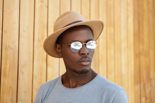 Concept de personnes, de style et de mode. À la mode à la mode jeune mannequin afro-américain portant des lunettes de soleil miroir et coiffe posant au mur en bois avec copie espace pour votre information