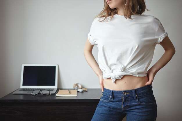 Concept de personnes, style, mode, vêtements et design. Belle jeune femme élégante avec un corps mince et élancé posant à l'intérieur avec un ordinateur portable en arrière-plan, vêtu d'un t-shirt blanc avec fond pour votre logo