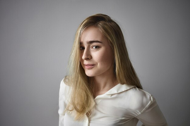 Concept de personnes et de style de vie. Belle jeune femme gestionnaire aux cheveux blonds raides et aux yeux verts posant en chemisier blanc au bureau, ayant un regard joyeux et heureux, se réjouissant des réalisations de carrière