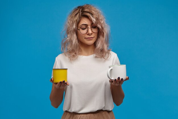 Concept de personnes, de style de vie, de boisson et de nourriture. Drôle de jeune femme douteuse indécise avec des cheveux roses en désordre face au dilemme, hésitant en choisissant entre le café et le thé