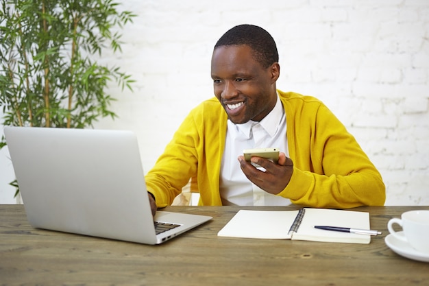 Concept de personnes, de style de vie, d'emploi, de technologie et de communication. Cheerful male pigiste afro-américain en cardigan jaune vif à l'aide de téléphone portable et ordinateur portable au bureau à domicile, souriant largement