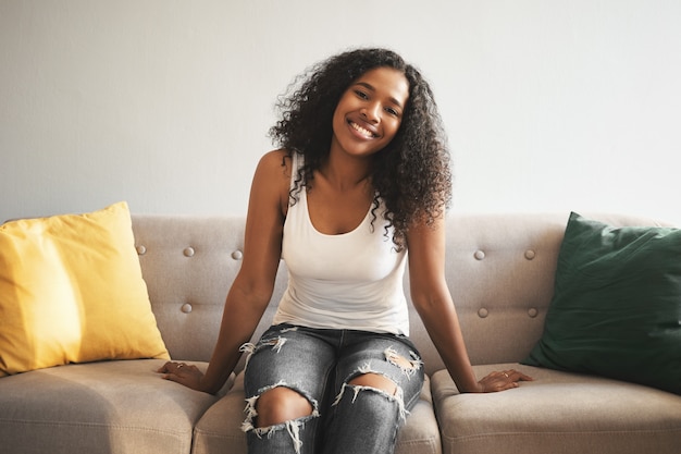 Concept de personnes, style de vie, loisirs, repos et relaxation. Adorable belle jeune femme à la peau sombre portant un débardeur blanc et un jean déchiré ayant un regard insouciant heureux tout en vous relaxant à la maison sur le canapé