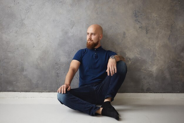 Concept de personnes, de style de vie, de repos et de relaxation. Beau jeune homme mal rasé à la mode avec tête chauve et chaume assis sur le sol de son nouvel appartement, ayant un regard pensif pensif, rêverie