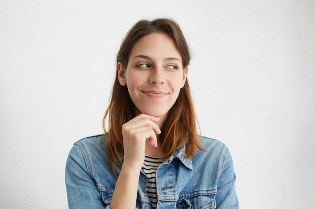 Concept de personnes et de style de vie. Tir horizontal de jolie fille avec un charmant sourire énigmatique, tenant la main sur son menton et en détournant les yeux