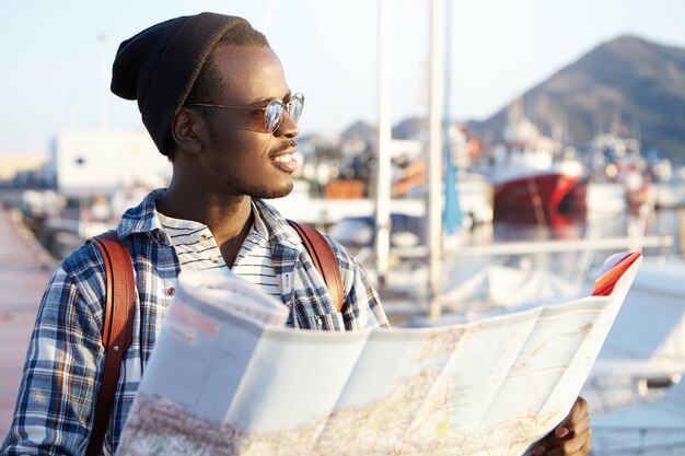 Concept de personnes, de style de vie, de voyages et d'aventures. L'homme sur le quai entouré de yachts et de navires à la mode chapeau et lunettes de soleil miroir tenant guide papier à côté en mer avec sourire heureux