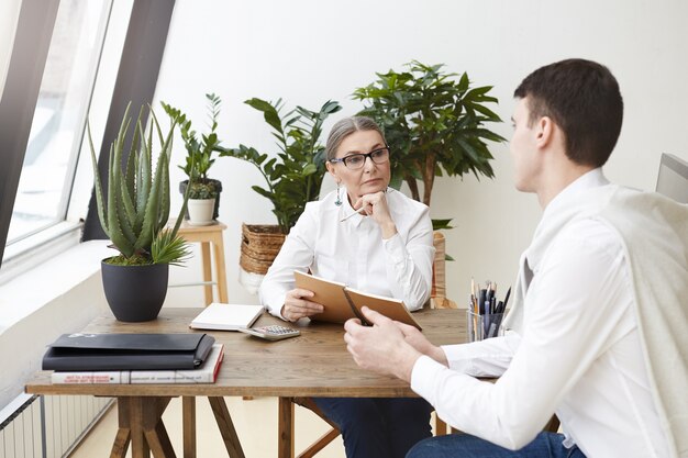 Concept de personnes, de travail, d'emploi, de profession et de profession. Expérimenté sérieux aux cheveux gris femme mature spécialiste des ressources humaines tenant un cahier tout en menant un entretien d'embauche avec un jeune homme