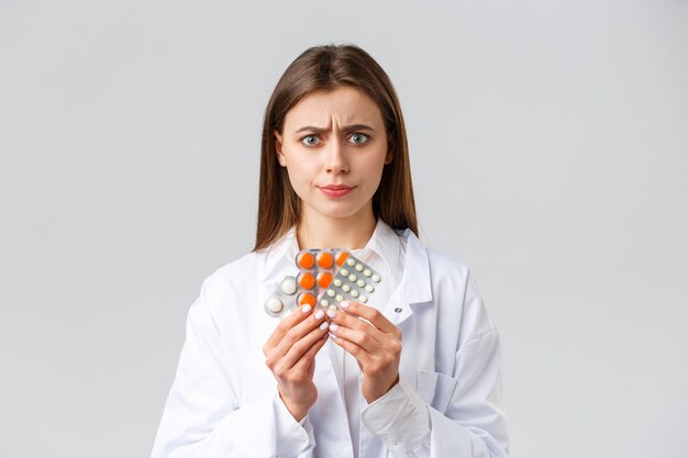 Concept de pharmacie, de travailleurs de la santé, d'assurance et d'hôpitaux. Femme médecin inquiète et sceptique en gommage blanc, a l'air méfiante, n'aime pas ce médicament, sourit déçue, montre un mauvais médicament