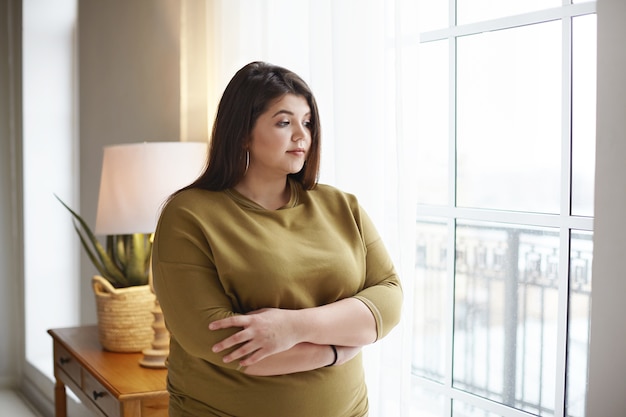 Concept de positivité de personnes, de mode de vie et de corps. Belle jeune femme brune avec des kilos en trop debout près de la fenêtre à la maison, croisant les bras sur sa poitrine, ayant un regard pensif, réfléchissant